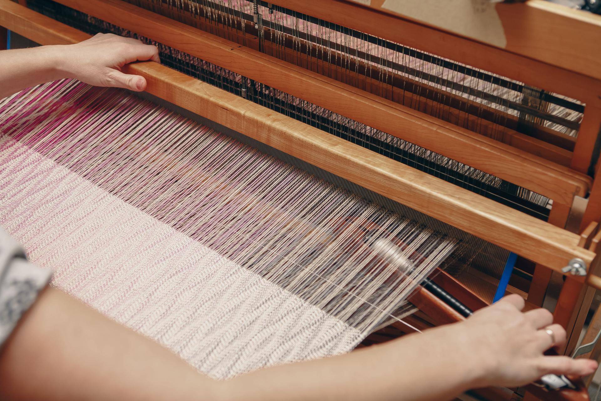 Hands operating a loom while weaving a light-colored textile.