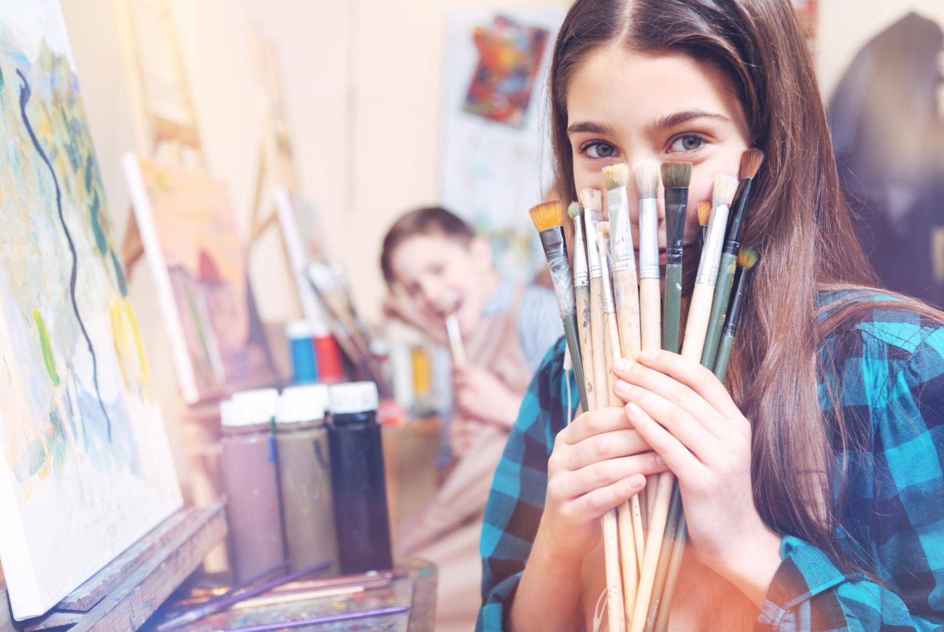 Child holding paintbrushes in an art classroom while another child paints in the background.
